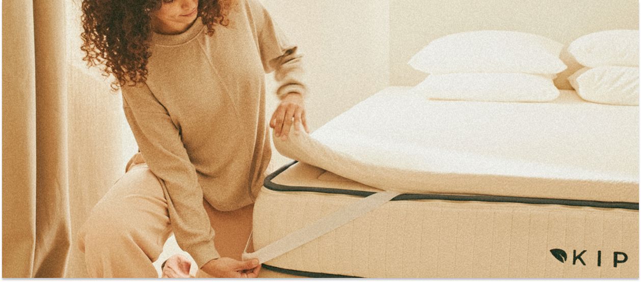 Eine Frau legt eine Matratzenauflage auf eine Kipli-Matratze aus Naturlatex in einem hellen, minimalistisch eingerichteten Schlafzimmer.