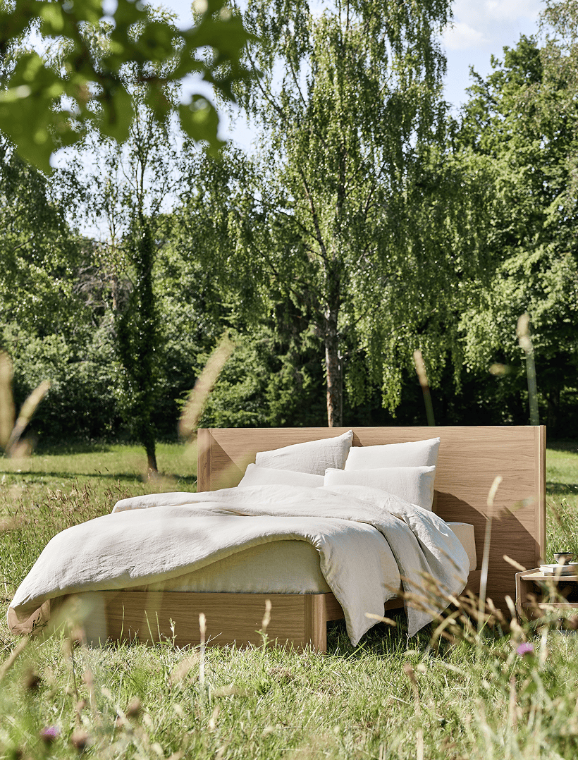 Ein Kipli-Bett aus Massivholz inmitten der Natur, mit Bettwäsche aus Naturleinen – ein Beispiel für ein umweltfreundliches Schlafzimmer und gesunden Schlaf inmitten einer grünen Umgebung.