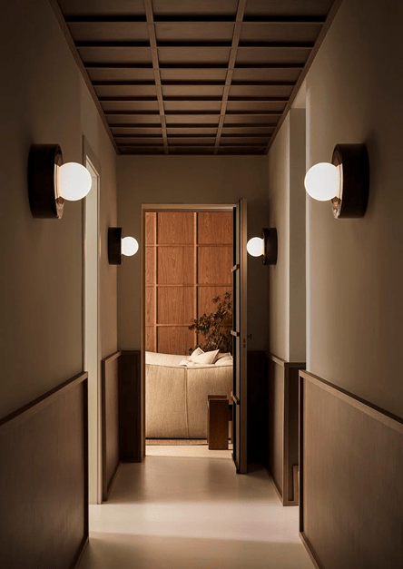 Couloir minimaliste éclairé par des appliques murales modernes, menant à un salon chaleureux en bois et canapé beige, illustrant une décoration intérieure élégante et apaisante.