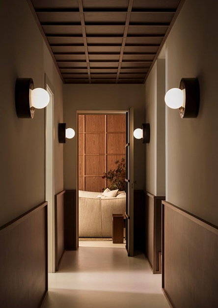Ein minimalistischer Flur, beleuchtet von modernen Wandleuchten, der zu einem gemütlichen Wohnzimmer mit Holzmöbeln und einem beigen Sofa führt und eine elegante und beruhigende Inneneinrichtung verkörpert.