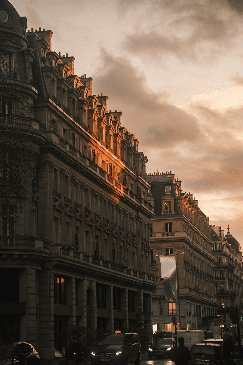 Hausmann-Fassaden in Paris bei Sonnenuntergang, mit goldenem Licht auf den Dächern und Verkehr vor den Gebäuden.