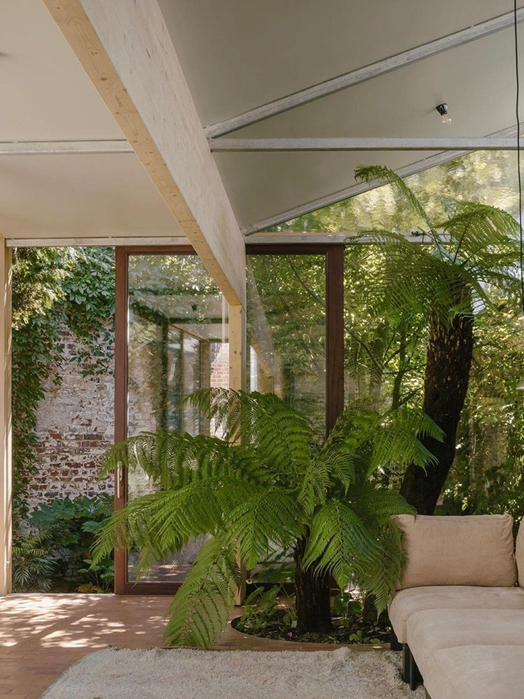 Ein heller Wohnbereich mit natürlicher Einrichtung, großen Grünpflanzen und einer Fensterfront mit Blick auf den Garten, der ein beruhigendes und umweltbewusstes Wohnambiente vermittelt.