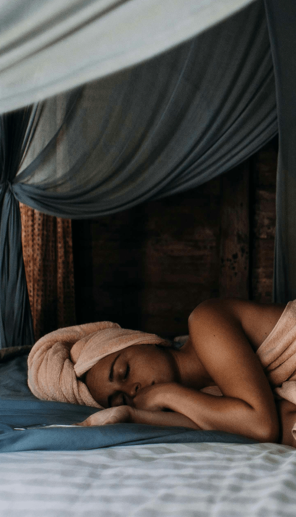 Femme se reposant après un moment de détente, allongée sur un lit entouré de rideaux drapés, illustrant un sommeil apaisant et une ambiance bien-être naturelle.