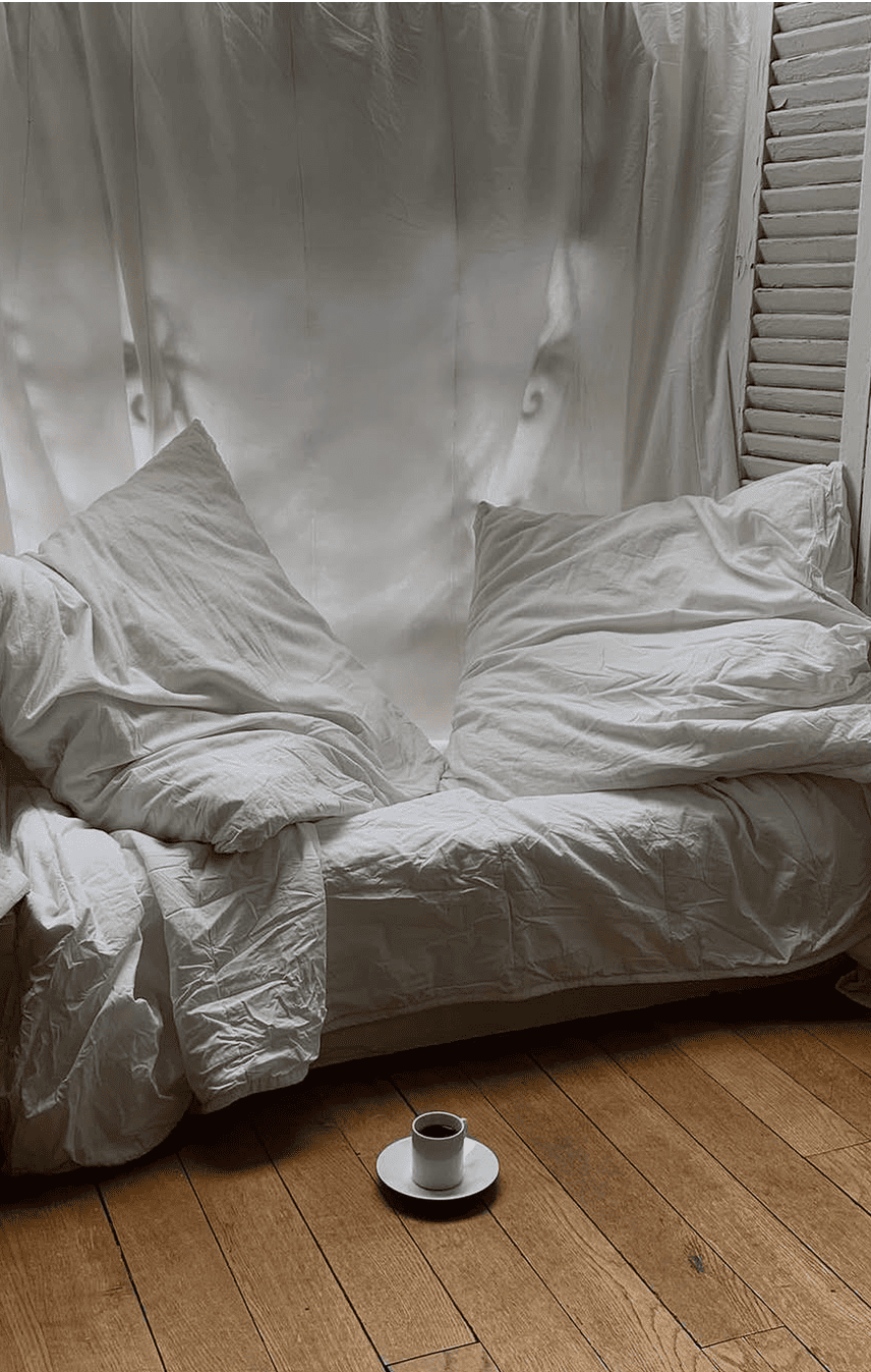 Ein gemütliches Schlafzimmer mit ungemachtem Bett in weißer Bettwäsche und einer auf dem Boden stehenden Kaffeetasse, das eine „Slow Living“-Atmosphäre und ein natürliches Erwachen vermittelt.