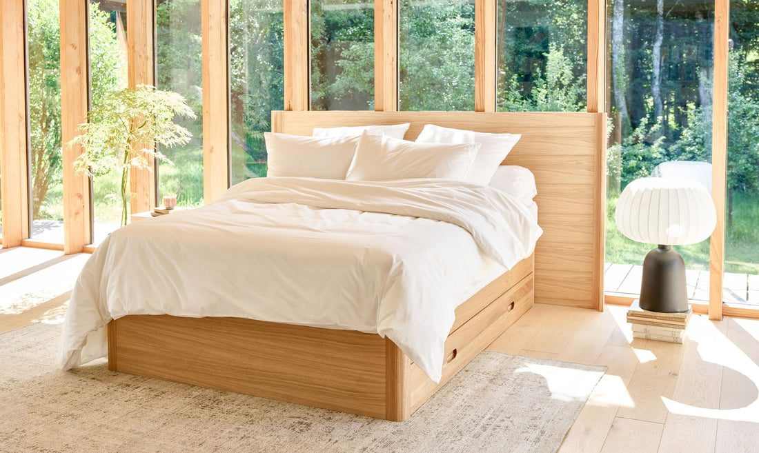Ein helles Zimmer mit einem Kipli-Bett aus Eichenholz mit Schubladen, weißer Bettwäsche und Tageslicht, das durch große Fensterfronten hereinströmt.