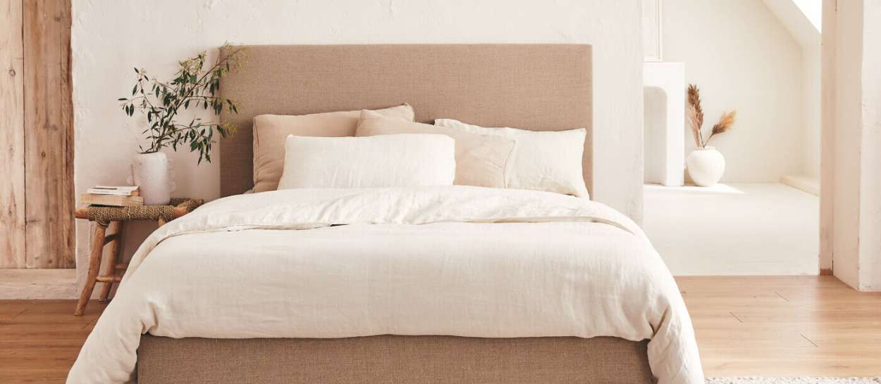Ein Kipli-Bett mit beigem Stoffbezug und heller Bettwäsche in einem hellen, natürlichen Schlafzimmer