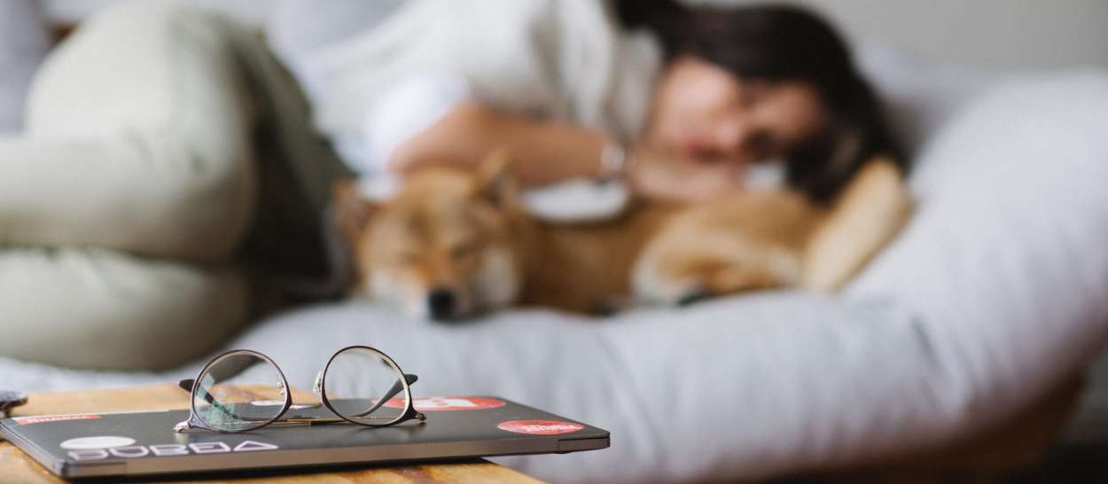Eine Frau, die sich mit ihrem Hund auf dem Bett ausruht, während ihre Brille auf dem Tisch liegt